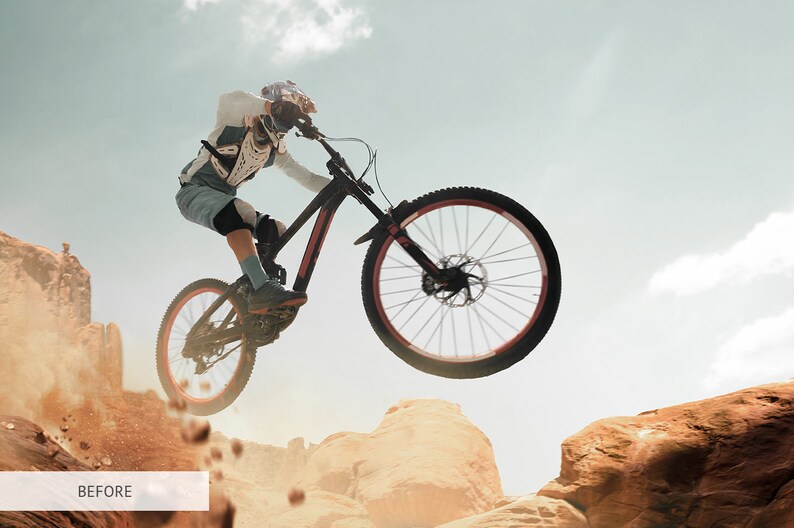 May include: A person wearing protective gear rides a black mountain bike over a rocky terrain. The bike is in mid-air, with the front wheel off the ground. The rider is wearing a helmet, goggles, and a long-sleeved shirt. The background is a blue sky with white clouds.