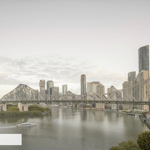 May include: A view of a large bridge over a river with a city skyline in the background. The bridge is made of steel and has a gray color. The city skyline is made up of tall buildings, some of which are skyscrapers. The river is wide and calm, and the sky is a light gray color.