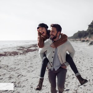 May include: A couple is walking on a sandy beach. The man is carrying the woman on his back. They are both smiling and laughing. The woman is wearing a brown sweater and a black hat. The man is wearing a white sweater and gray pants.