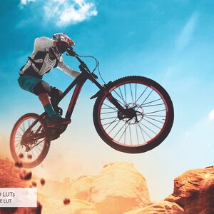 May include: A mountain biker in mid-air, performing a jump over a rocky terrain. The biker is wearing a helmet and protective gear, and the bike is black with red accents. The sky is blue with white clouds, and the ground is a mix of red dirt and rocks.