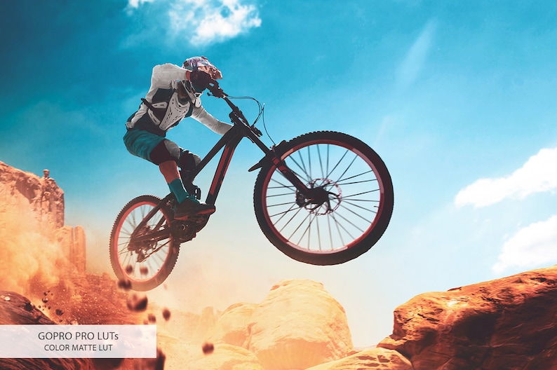 May include: A mountain biker in mid-air, performing a jump over a rocky terrain. The biker is wearing a helmet and protective gear, and the bike is black with red accents. The sky is blue with white clouds, and the ground is a mix of red dirt and rocks.