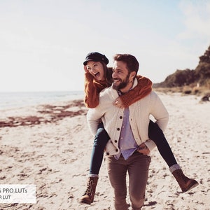 May include: A couple laughing and walking on a sandy beach. The man is carrying the woman on his back. They are both wearing casual winter clothing.