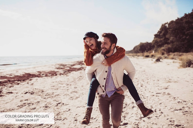 May include: A couple laughing and walking on a sandy beach. The man is carrying the woman on his back. They are both wearing casual winter clothing.