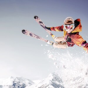 May include: A skier in a red and yellow jacket and goggles jumps in the air with skis extended. The skier is against a blue sky with snow-covered mountains in the background.