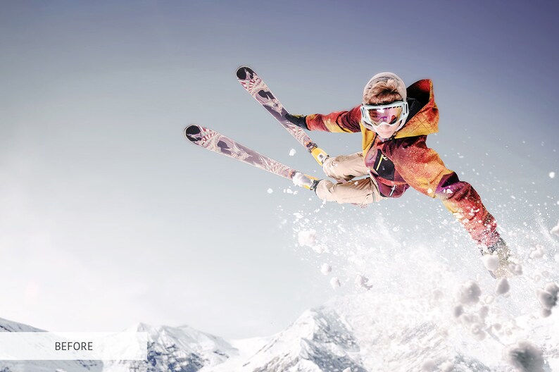 May include: A skier in a red and yellow jacket and goggles jumps in the air with skis extended. The skier is against a blue sky with snow-covered mountains in the background.