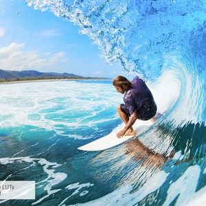 May include: A surfer rides a wave, crouching on a surfboard, with a blue sky and green mountains in the background.