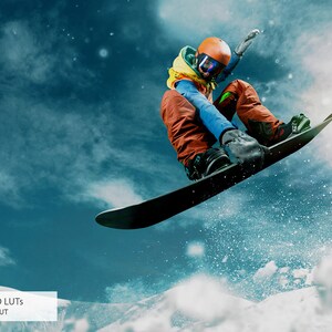 May include: A snowboarder wearing a blue jacket, orange pants, and a yellow helmet jumps in the air against a blue sky with white clouds. The snowboarder is performing a trick with their arms outstretched. The snowboard is black and has a white base.