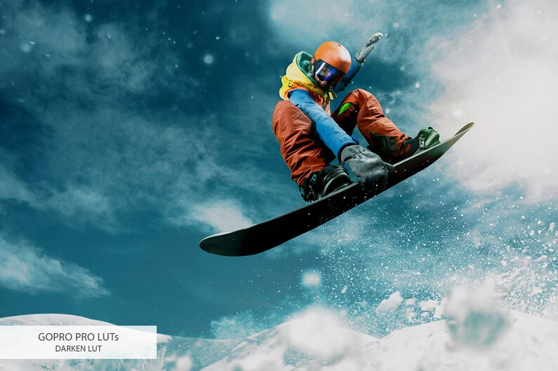 May include: A snowboarder wearing a blue jacket, orange pants, and a yellow helmet jumps in the air against a blue sky with white clouds. The snowboarder is performing a trick with their arms outstretched. The snowboard is black and has a white base.