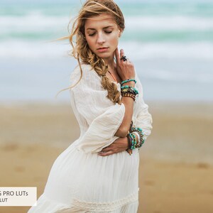 May include: A woman with long blonde hair wearing a white dress and multiple turquoise and brown bracelets stands on a sandy beach with the ocean in the background.