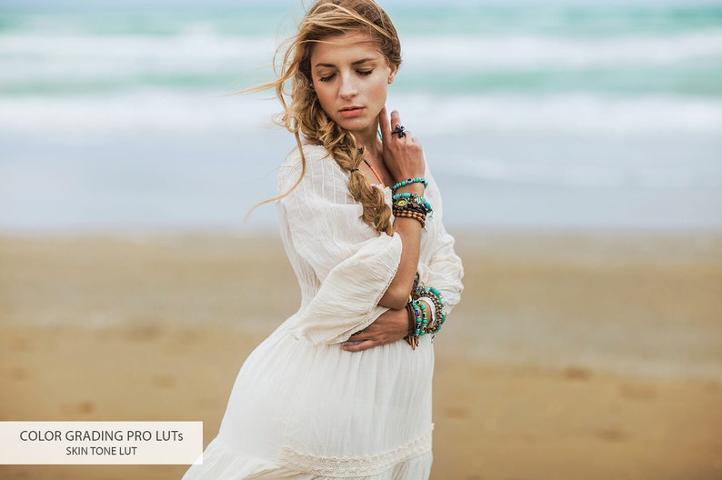 May include: A woman with long blonde hair wearing a white dress and multiple turquoise and brown bracelets stands on a sandy beach with the ocean in the background.