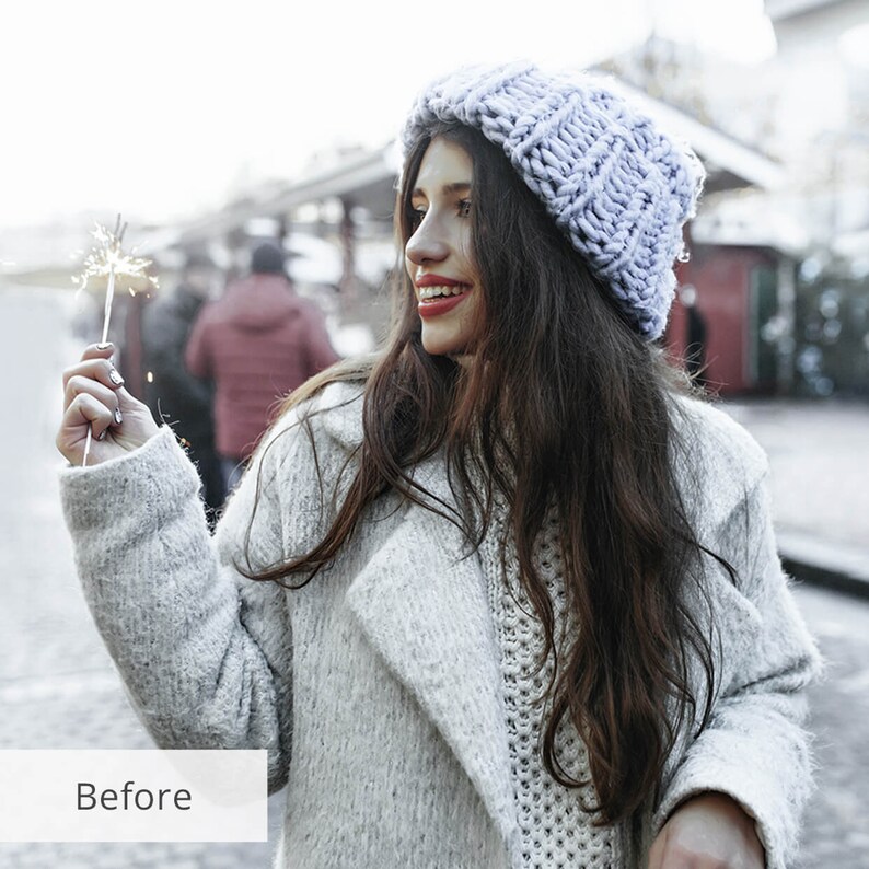 May include: A woman wearing a light gray knitted coat and a light purple knitted beanie holds a silver sparkler in her right hand. She is smiling and looking to the right. The background is blurred and shows a city street.