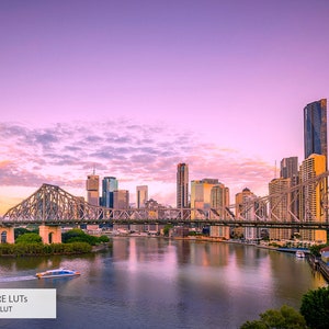 May include: A panoramic view of a city skyline at sunset with a bridge crossing a river. The sky is a vibrant pink and purple with fluffy clouds. The city buildings are tall and modern, with a few skyscrapers visible. The bridge is made of steel and has a distinctive arch design. The river is calm and reflective, reflecting the city lights.