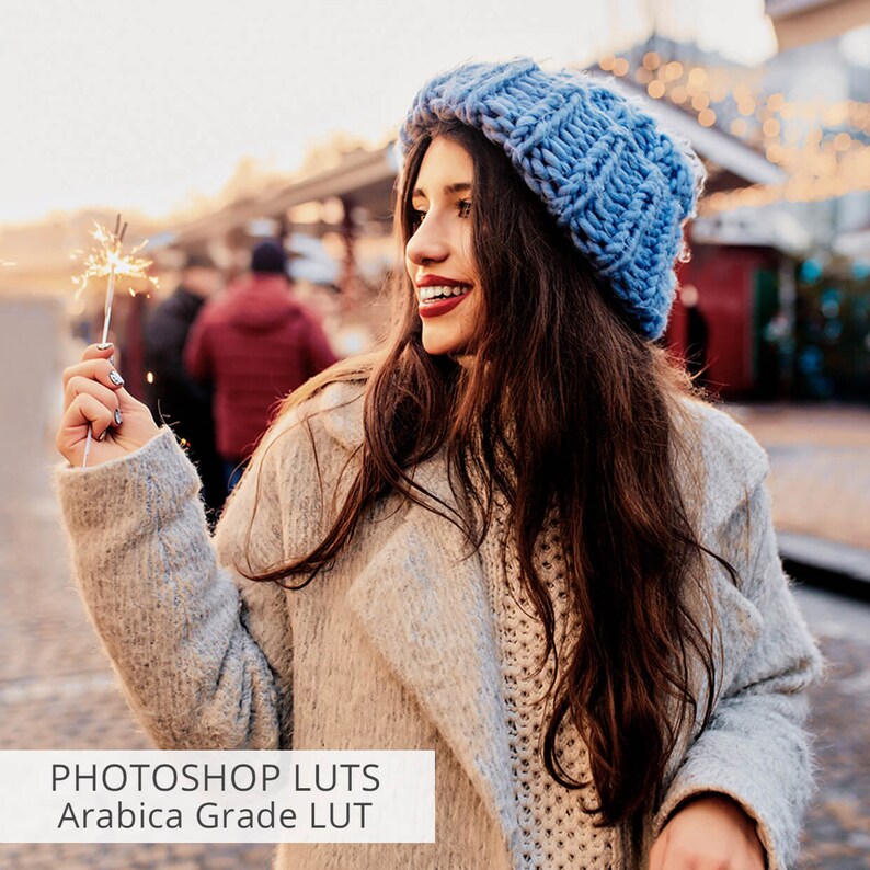 May include: A woman wearing a blue knitted hat and a beige coat holds a lit sparkler in her hand. The background is a blurred image of a city street with Christmas lights.