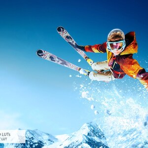 May include: A skier in a red, yellow, and blue jacket jumps through the air with skis extended. The skier is wearing a helmet and goggles. The background is a blue sky with snow-covered mountains.