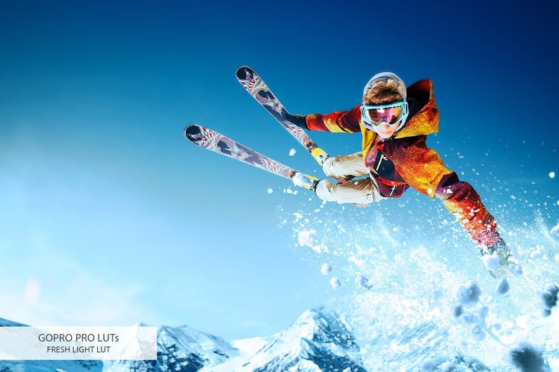 May include: A skier in a red, yellow, and blue jacket jumps through the air with skis extended. The skier is wearing a helmet and goggles. The background is a blue sky with snow-covered mountains.