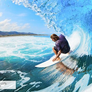 May include: A surfer rides a wave, crouching on a surfboard, with a blue sky and green mountains in the background.
