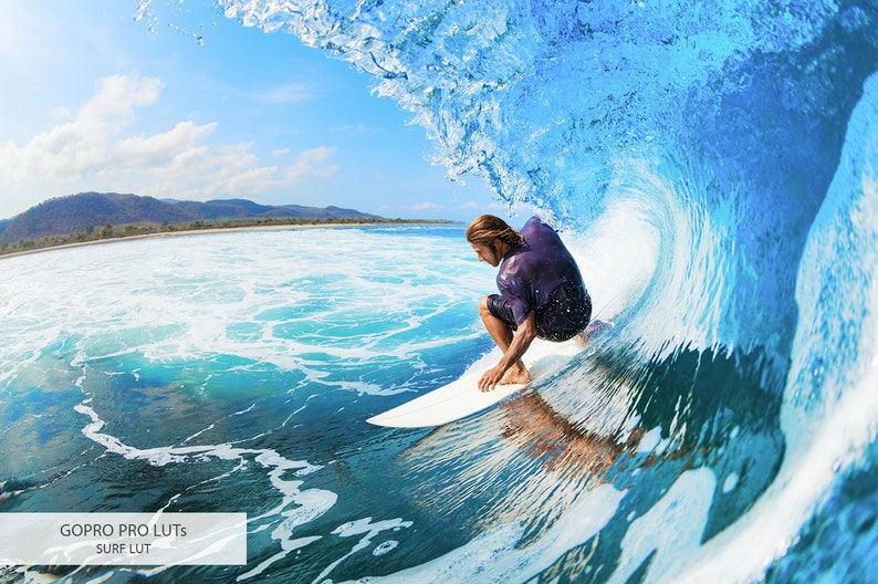 May include: A surfer rides a wave, crouching on a surfboard, with a blue sky and green mountains in the background.