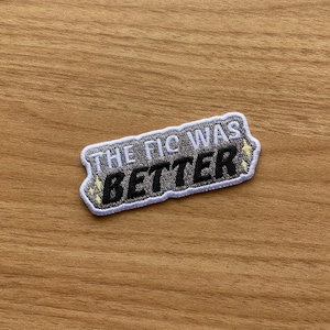 Puede incluir: Parche bordado con el texto "THE FIC WAS BETTER" en blanco y negro, con un borde gris. La palabra "BETTER" está en negrita, con una fuente estilizada. El parche tiene acentos de estrellas amarillas.