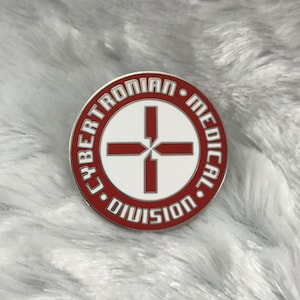 May include: A red and white enamel pin with the text "Cybertronian Medical Division" in a circle around a red cross.