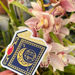May include: A blue and gold sticker of a closed book with a crescent moon and a symbol on the cover. The sticker is on a pink orchid flower.