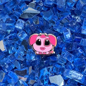 May include: A metal enamel pin shaped like a cartoon pig with a light pink face, dark pink ears, and large black eyes. The pig has a small snout and a tiny tail, with a metallic gold outline. The pin is resting on a bed of blue glass shards.