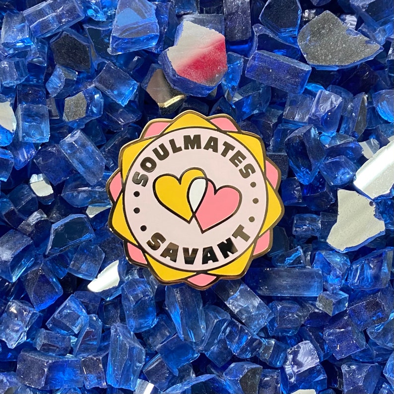 May include: A gold-colored pin with a pink and yellow heart design. The pin says "Soulmates Savant" in a circle around the hearts.