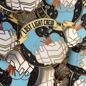 May include: A close-up of a gold and white enamel pin with a blue and white cloud design. The pin features the text "LOST LIGHT CREW" on a yellow banner.