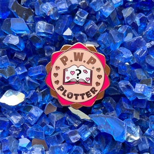 May include: A pink enamel pin with a white outline and the text "P.W.P. PLOTTER" in a circle. The pin features a cartoon illustration of a pink book with a question mark and hearts.
