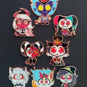 May include: Set of nine enamel pins featuring characters from the anime series Helluva Boss. The pins are colorful and feature various characters, including a red demon with horns, a blue demon with a top hat, and a white cat with a pink bow.