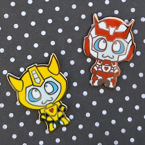 May include: Two enamel pins featuring cartoon robot characters. The first pin is yellow and features a robot with large blue eyes. The second pin is red and features a robot with large white eyes.