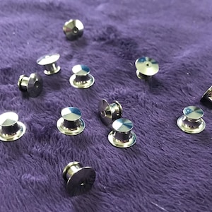 May include: A close-up of twelve silver metal pin backs on a purple fuzzy surface.