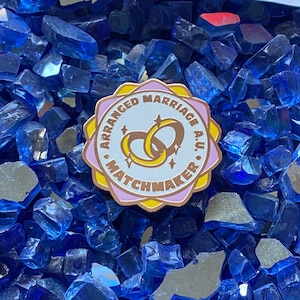 May include: A pink and gold enamel pin with the text "Arranged Marriage A.U. Matchmaker" and two interlocking rings with stars inside.