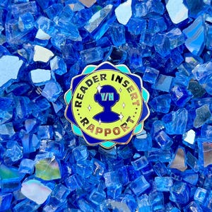 May include: A blue and yellow enamel pin with the text "...READER INSERT RAPPORT..." and a stylized alien head with the text "V/N" inside. The pin is on a blue background.