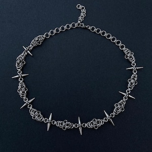 Op de afbeelding: Een zilveren ketting met scherpe, puntige spikes. De ketting is gemaakt van kleine, met elkaar verbonden metalen schakels.