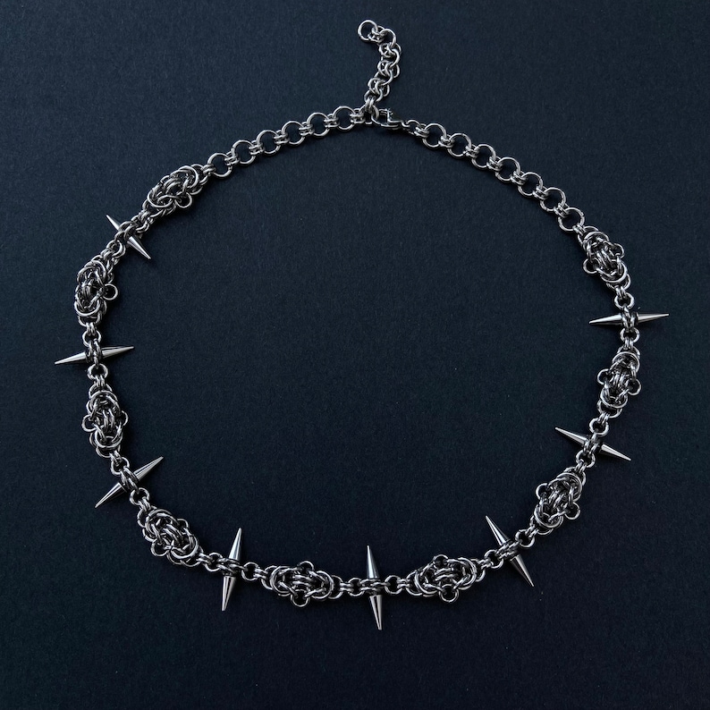 Op de afbeelding: Een zilveren ketting met scherpe, puntige spikes. De ketting is gemaakt van kleine, met elkaar verbonden metalen schakels.