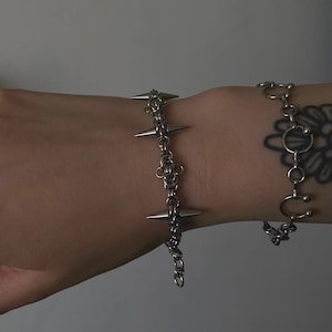 May include: Two silver bracelets on a person's wrist. One bracelet is a chain with spikes, the other is a chain with a circular charm.