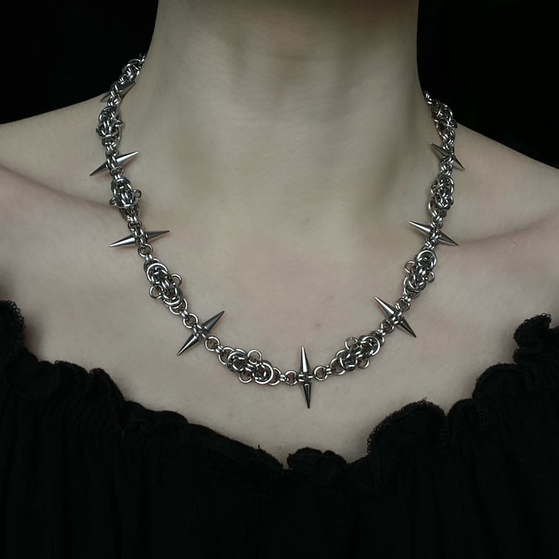 Op de afbeelding: Een zilveren ketting met scherpe, puntige spikes. De ketting is gemaakt van een geweven metalen ketting en heeft in totaal acht spikes.