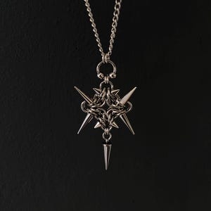 Peut inclure: Collier pendentif argenté avec un motif géométrique et des éléments pointus. Le pendentif est suspendu à une chaîne délicate. Le collier a une esthétique gothique ou edgy, sur fond sombre.