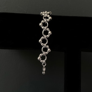 May include: A silver-toned chain bracelet featuring a repeating pattern of small metallic spheres and circular links. The design consists of alternating segments of round beads and open rings, finished with a small lobster claw clasp at the end.