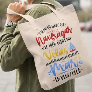May include: A white canvas tote bag with a colourful graphic design. The text on the bag reads "Y SI UN DIA TENGO QUE. Naufragar Y EL TIF&Oacute;N ROMPE MIS Velas ENTERRAD MI CUERPO CERCA DEL Mar EN VENEZUELA."