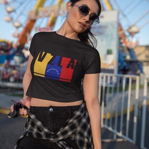 May include: A black crop top with a graphic of the Venezuelan flag and the letters "VZL" in red, yellow, and blue. The model is wearing black denim shorts and a black and white plaid shirt tied around her waist.