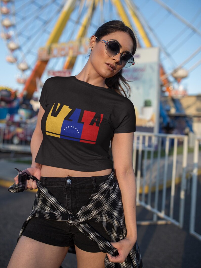 May include: A black crop top with a graphic of the Venezuelan flag and the letters "VZL" in red, yellow, and blue. The model is wearing black denim shorts and a black and white plaid shirt tied around her waist.