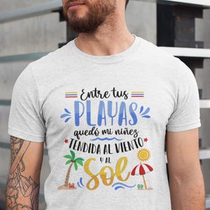 May include: White t-shirt with a graphic design featuring the text "Entre tus playas qued&oacute; mi ni&ntilde;ez tendida al viento y al sol". The graphic also includes a palm tree, a beach umbrella, and a crab.