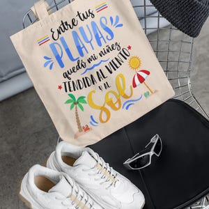 May include: A beige canvas tote bag with the text "= Entre tus = PLAYAS qued&oacute; mi ni&ntilde;ez *TENDIDA AL VIENTO y al Sol" in colourful lettering. The bag is sitting on a black chair with a grey beanie and white trainers.