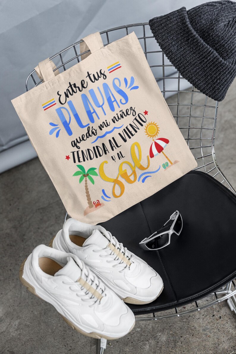 May include: A beige canvas tote bag with the text "= Entre tus = PLAYAS qued&oacute; mi ni&ntilde;ez *TENDIDA AL VIENTO y al Sol" in colourful lettering. The bag is sitting on a black chair with a grey beanie and white trainers.