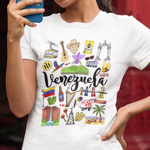 May include: White t-shirt with a vibrant illustration of Venezuela. The design includes a cable car, guitar, a man in a hat, a parrot, a mountain, a bus, a flag, a waterfall, and the word "Venezuela" in cursive.