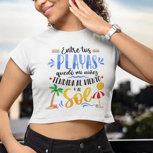 May include: White crop top with the text "Entre tus playas qued&oacute; mi ni&ntilde;ez tendida al viento y al sol" with a palm tree and an umbrella graphic.