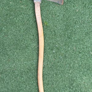 May include: A hand tool featuring a long, curved wooden handle attached to a metal head with a single, wide, curved blade. The metal head is dark gray with signs of wear, and the wooden handle has a smooth, light brown finish.
