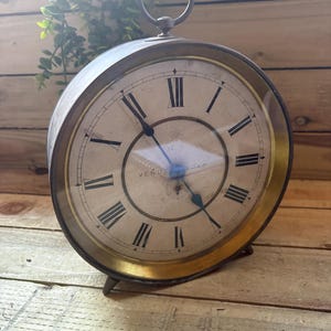 May include: A vintage-style clock with a circular face and Roman numerals. The clock features a gold-coloured metal frame and a handle at the top. The clock face has the words "Venus Soap" printed on it. The clock is on a wooden surface.