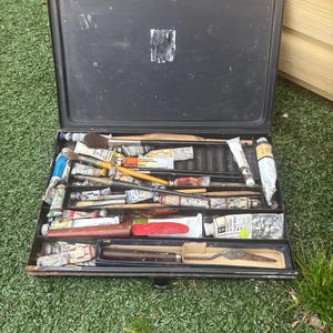 Pode incluir: Uma caixa de pintura preta aberta, cheia de vários tubos de tinta, pincéis e uma espátula. Os tubos de tinta são de várias cores, e os pincéis têm cabos de madeira. A caixa está sobre a relva verde.